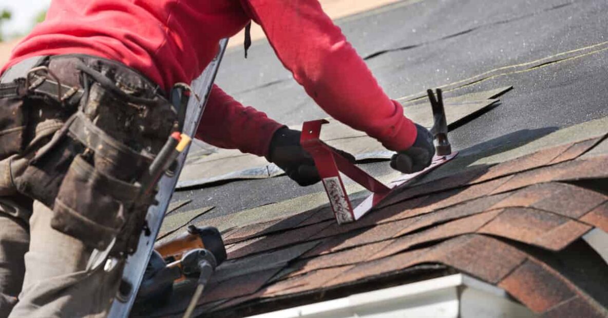 A person in a red sweatshirt and work pants is working on a roof, using tools to install or repair shingles.