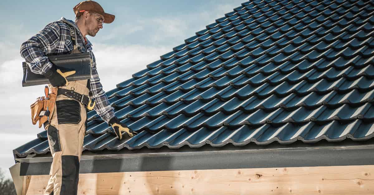 A worker in a plaid shirt and work pants is inspecting a dark-colored tiled roof.